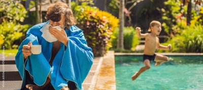 Mann sitzt im Bademantel am Rand eines Swimming-Pools und putzt sich die Nase.