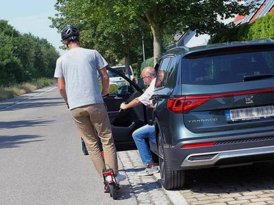 Ein Mann auf einem Scooter fährt an einem parkenden Auto vorbei, bei dem sich die Fahrertür öffnet.