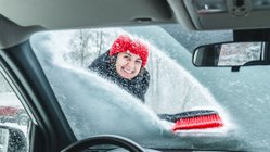 Junge Frau in Winterlandschaft befreit Autofrontscheibe von Schnee.